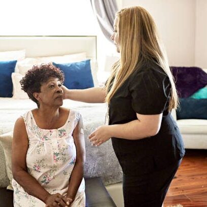 A caregiver comforts an elderly woman sitting on a chair in a cozy living room.